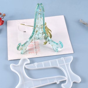 Könnte beinhalten: Zwei transparente Harz-Präsentationsständer mit goldenen Flocken. Die Ständer haben die Form eines Dreiecks mit abgerundeter Basis. Die Ständer stehen auf einer weißen Oberfläche.