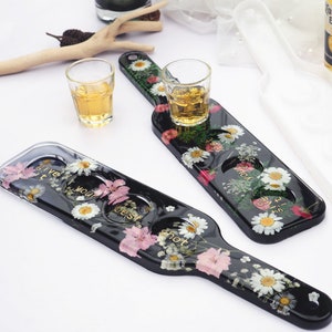 May include: Two black shot glass trays with floral designs and gold lettering. The trays have three shot glasses each. The trays are on a white surface. The text on the trays reads "Give it your best shot" and "Live your best life".
