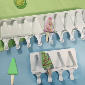 Może przedstawiać: Dwa białe plastikowe foremki do robienia lodów w kształcie choinki. Jedna foremka ma osiem otworów, a druga cztery. Na zdjęciu są trzy lody, jeden różowy z białymi smugami, jeden zielony i jeden czerwony z białymi smugami.