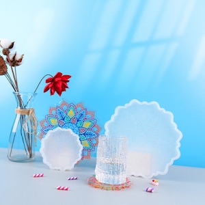 May include: A collection of decorative resin coasters in various sizes and designs. One coaster features a colorful mandala pattern, while others are clear with a floral design. A glass of water and a vase with flowers are also present.