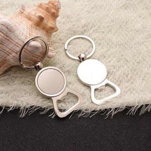 May include: Two silver bottle opener keychains with a round blank metal surface. The keychains are on a white fluffy surface.
