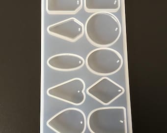 May include: A clear silicone mold with eight different shapes for making jewelry pendants. The shapes include a rectangle, a circle, an oval, a heart, a diamond, and three different teardrop shapes. Each shape has a small hole at the top for attaching a jump ring.