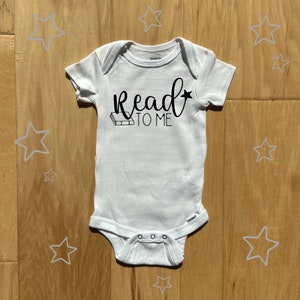 May include: White baby onesie with black text that reads "Read to Me" and a small illustration of a book.