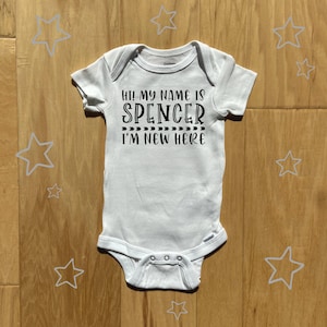 May include: White baby onesie with black text that reads "Hi! My name is Spencer I'm new here".