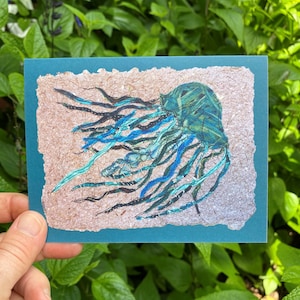May include: A teal greeting card featuring a detailed illustration of a jellyfish. The jellyfish is depicted in shades of teal, blue, and black, with long, flowing tentacles. The artwork is set against a textured, light brown background, framed by a teal border.