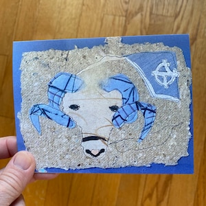 May include: A handmade greeting card with a ram's head illustration. The ram has blue horns and is set against a textured, beige background with a blue border. A blue flag with a white emblem is in the upper right corner.