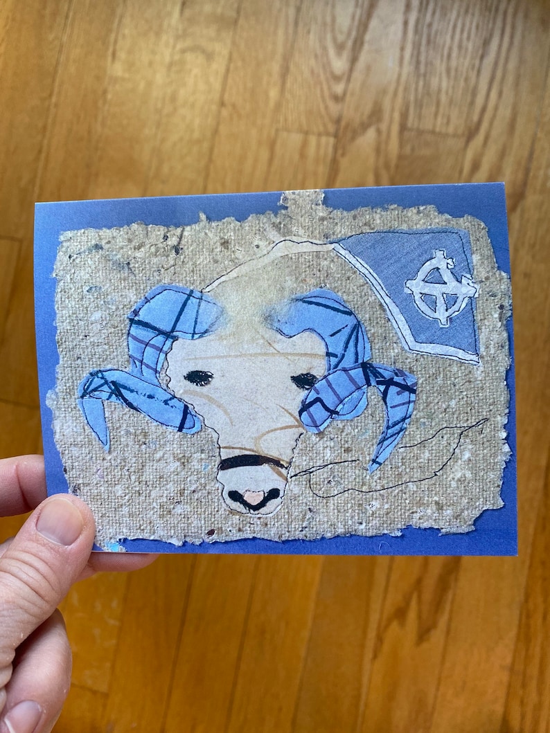 May include: A handmade greeting card with a ram's head illustration. The ram has blue horns and is set against a textured, beige background with a blue border. A blue flag with a white emblem is in the upper right corner.