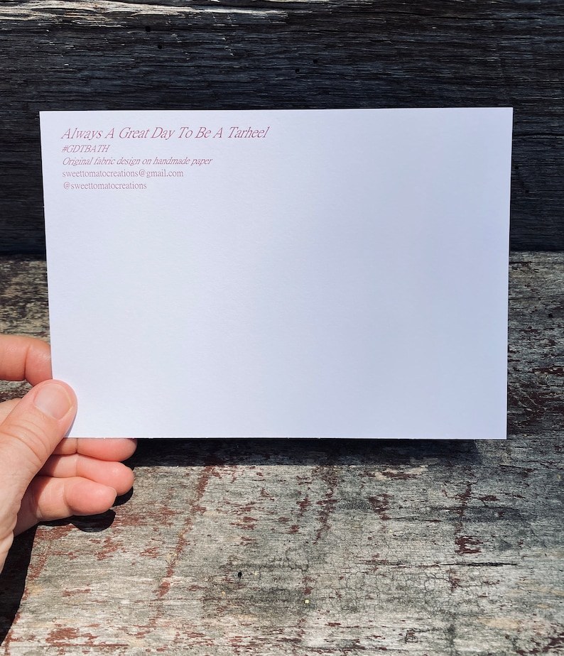 May include: A white card with the text "Always A Great Day To Be A Tarheel" in maroon. Additional text includes "#GDTBATH", and contact information for sweettomatocreations. The card is held against a weathered wooden surface.
