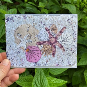 Peut inclure: Une carte faite à la main avec un collage de coquillages et d'étoiles de mer. La carte a un fond texturé et moucheté avec une variété de créatures marines dans des tons de rose, beige et blanc. La carte est tenue devant un fond de feuillage vert.