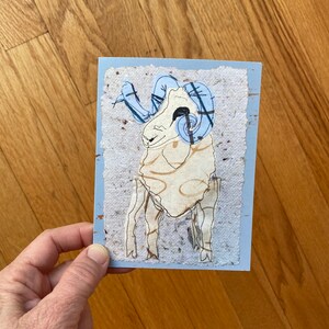 May include: A handmade greeting card featuring a ram illustration. The ram has a cream-colored body, light blue horns, and a light blue border. The card is held by a person.