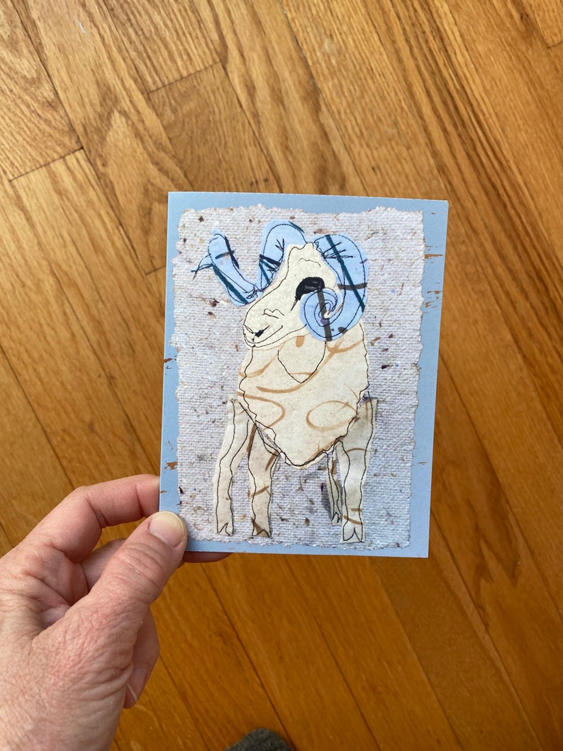 May include: A handmade greeting card featuring a ram illustration. The ram has a cream-colored body, light blue horns, and a light blue border. The card is held by a person.