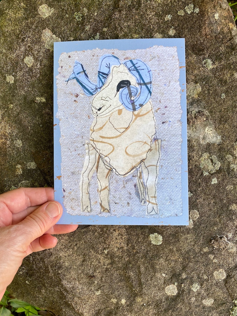 May include: A handmade greeting card featuring a ram illustration. The ram is depicted with a cream-colored body, black outlines, and blue horns. The card has a light blue border and a textured paper background.