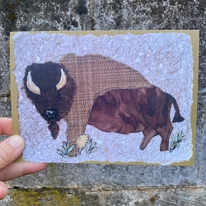 May include: A handmade bison art print on textured paper. The bison is depicted in shades of brown and tan, with a textured body and detailed features. The artwork is mounted on a light brown cardstock.