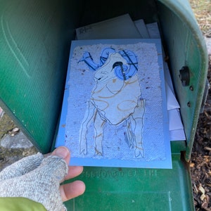 May include: A blue and cream-colored greeting card featuring a ram illustration with blue horns and gold accents. The card is inside a green mailbox, held by a gloved hand. The card has a textured paper appearance.