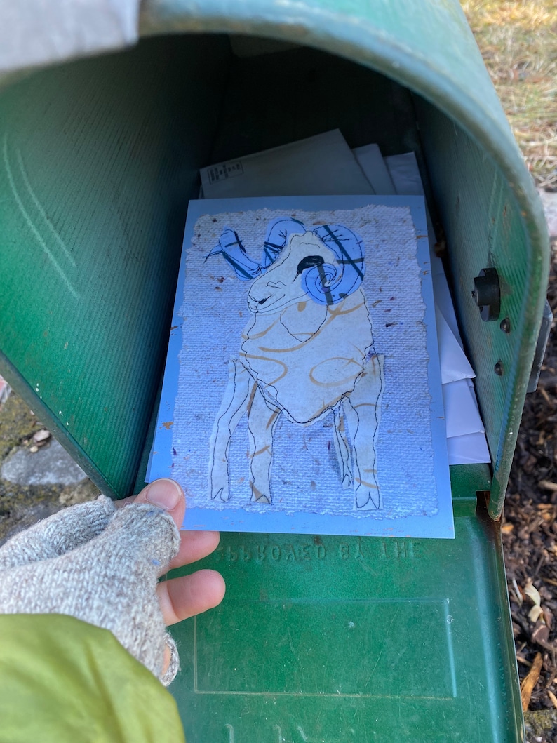 May include: A blue and cream-colored greeting card featuring a ram illustration with blue horns and gold accents. The card is inside a green mailbox, held by a gloved hand. The card has a textured paper appearance.