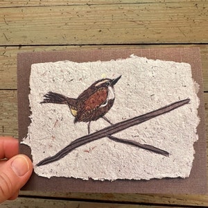 Puede incluir: Tarjeta de felicitación hecha a mano con una ilustración detallada de un pequeño pájaro posado en una rama. El pájaro tiene plumas marrones y blancas, con un acento amarillo. La tarjeta está sobre papel texturizado blanquecino con borde marrón.