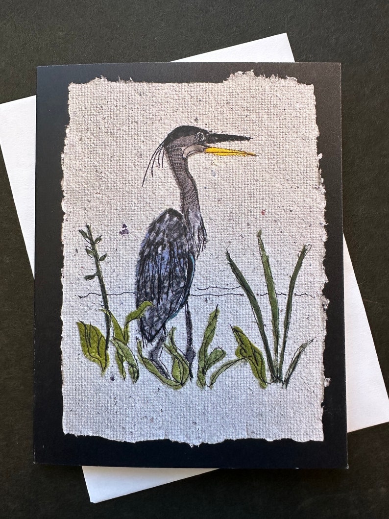 K&ouml;nnte beinhalten: Eine Gru&szlig;karte mit einer handgezeichneten Illustration eines Blaufischreihers in einem Sumpf. Der Reiher hat einen langen Hals und einen gelben Schnabel. Gr&uuml;ne Pflanzen umgeben den Vogel, und die Karte hat einen schwarzen Rand und einen wei&szlig;en Umschlag.