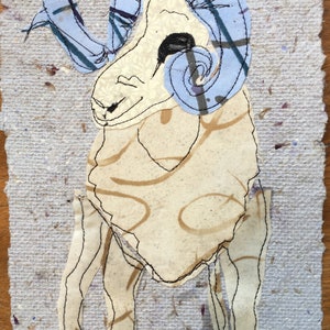 May include: A fabric art piece depicting a ram. The ram has a cream-colored body with brown swirls and blue horns with plaid accents. The artwork is stitched onto a textured, light gray paper background.