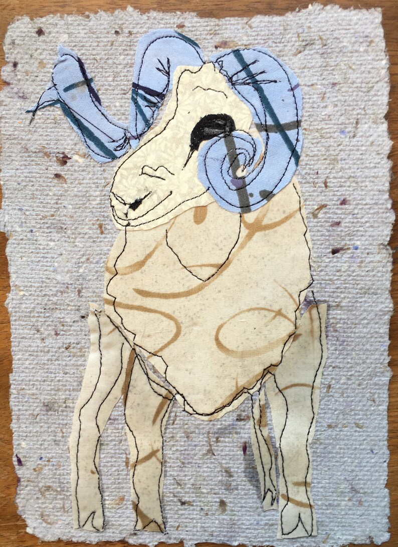 May include: A fabric art piece depicting a ram. The ram has a cream-colored body with brown swirls and blue horns with plaid accents. The artwork is stitched onto a textured, light gray paper background.