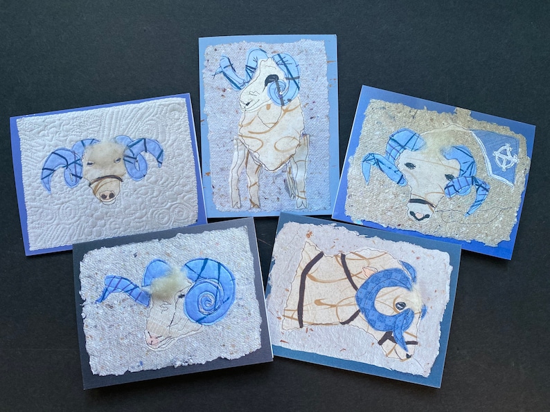 May include: Five handmade greeting cards with ram illustrations. Each card has a unique design with blue and tan accents, set against textured paper and blue borders. The ram's horns are a prominent feature.