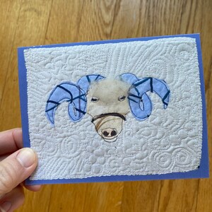 May include: A handmade greeting card depicting a ram's head. The ram has light blue horns and a tan face with a brown muzzle. The background is white with a quilted pattern, and the card has a blue border.