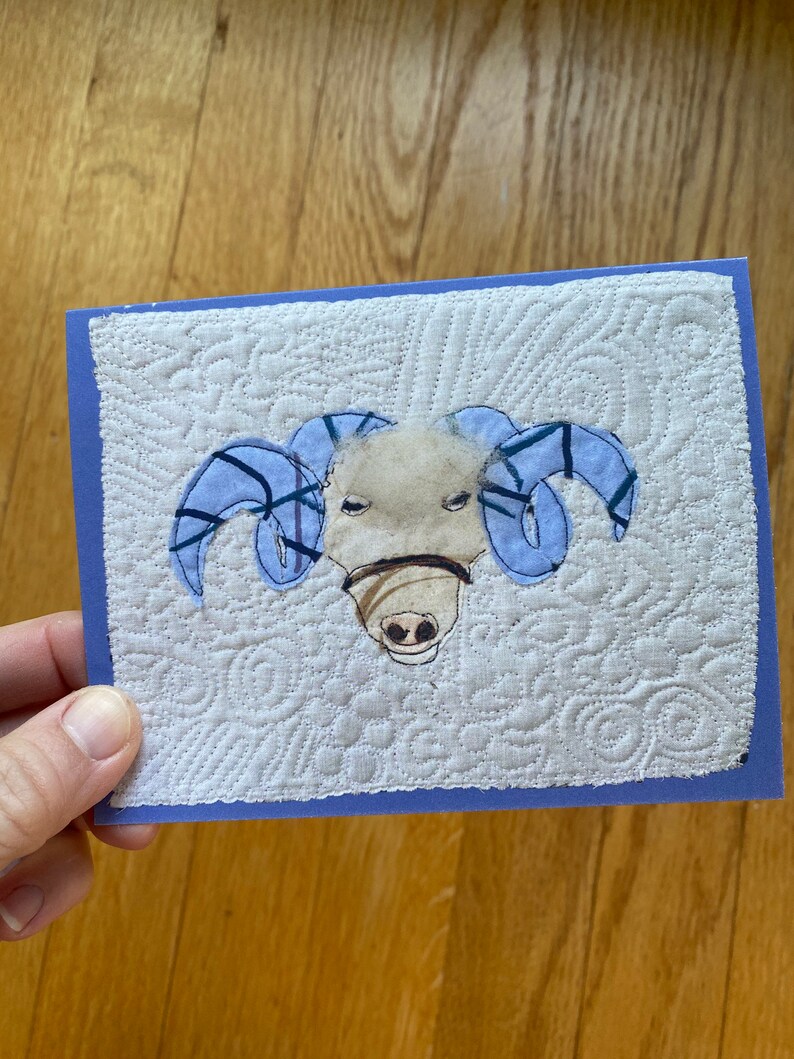 May include: A handmade greeting card depicting a ram's head. The ram has light blue horns and a tan face with a brown muzzle. The background is white with a quilted pattern, and the card has a blue border.