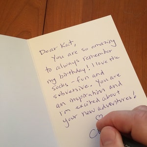 May include: A cream-coloured greeting card with a handwritten message in blue ink. The text reads, "Dear Kat, You are so amazing to always remember my birthday! I love the socks - fun and subversive. You are an inspiration and I'm excited about your new adventures!"