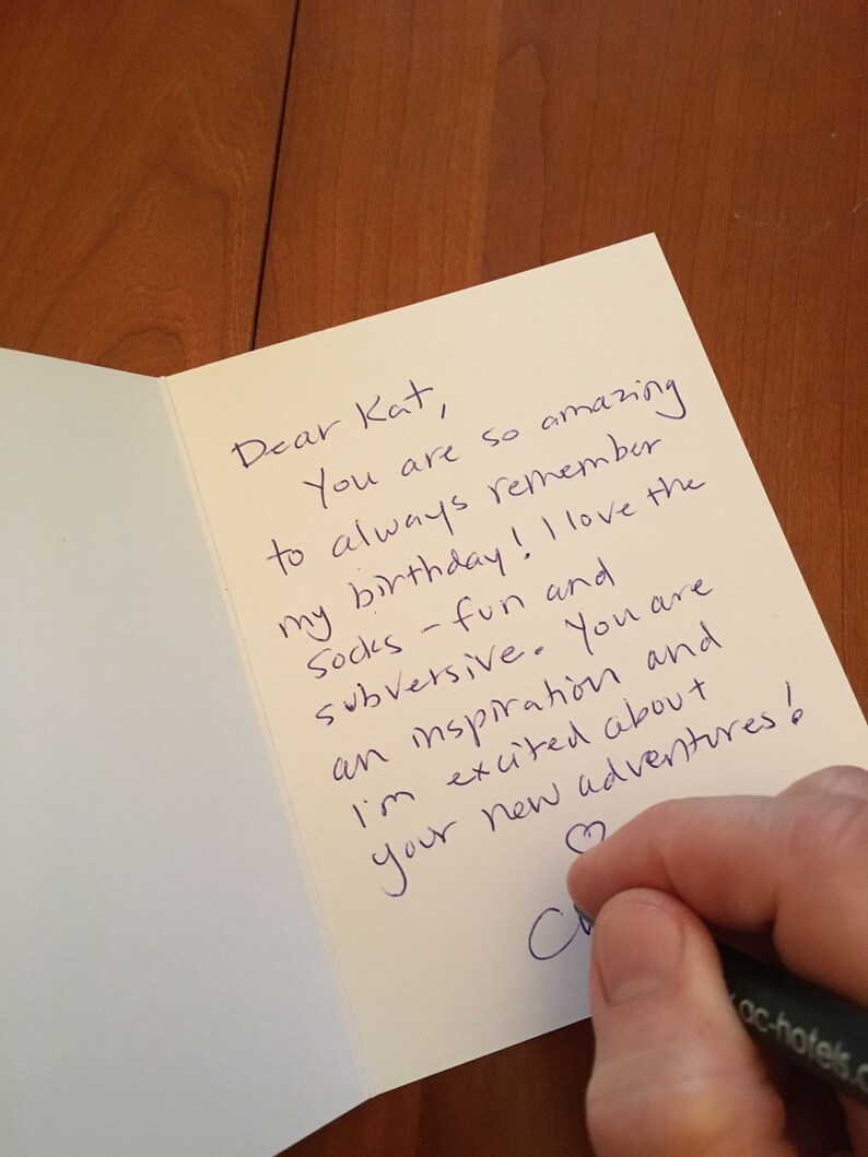 May include: A cream-coloured greeting card with a handwritten message in blue ink. The text reads, "Dear Kat, You are so amazing to always remember my birthday! I love the socks - fun and subversive. You are an inspiration and I'm excited about your new adventures!"
