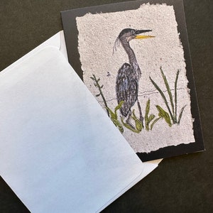 K&ouml;nnte beinhalten: Eine Gru&szlig;karte mit einer Illustration eines Fischreihers, der im Wasser steht, umgeben von gr&uuml;nen Schilf. Der Hintergrund ist strukturiert, cremefarben, mit einem schwarzen Rand. Ein wei&szlig;er Umschlag ist teilweise sichtbar.