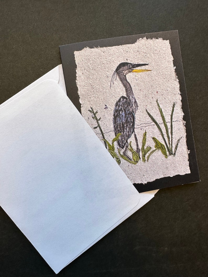K&ouml;nnte beinhalten: Eine Gru&szlig;karte mit einer Illustration eines Fischreihers, der im Wasser steht, umgeben von gr&uuml;nen Schilf. Der Hintergrund ist strukturiert, cremefarben, mit einem schwarzen Rand. Ein wei&szlig;er Umschlag ist teilweise sichtbar.