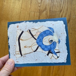 May include: A handmade greeting card with a ram illustration. The ram has a blue patterned scarf and horns, set against a textured, off-white background with brown accents. The card has a dark blue border.