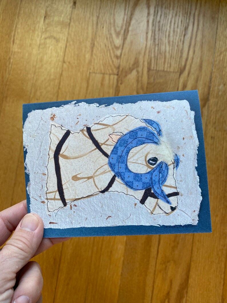 May include: A handmade greeting card with a ram illustration. The ram has a blue patterned scarf and horns, set against a textured, off-white background with brown accents. The card has a dark blue border.