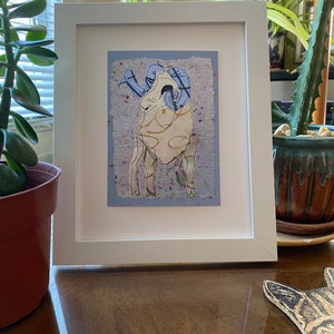 May include: White framed artwork featuring a stylized ram illustration. The ram has blue horns and is set against a textured, light blue background. The artwork is displayed on a wooden surface, with plants visible in the background.