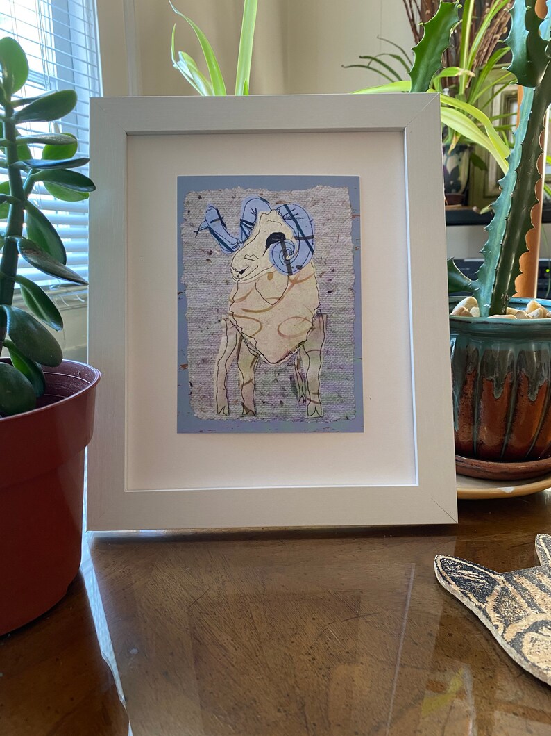 May include: White framed artwork featuring a stylized ram illustration. The ram has blue horns and is set against a textured, light blue background. The artwork is displayed on a wooden surface, with plants visible in the background.