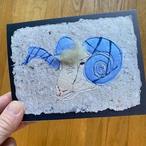 May include: A handmade greeting card with a ram's head applique. The ram has light blue horns and a cream-coloured face. The applique is on a textured, off-white background with a black border. The card is held by a hand.