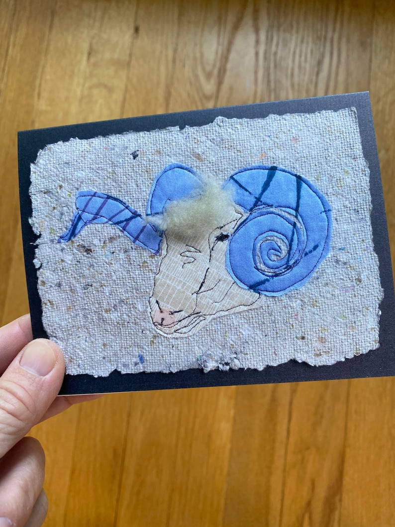 May include: A handmade greeting card with a ram's head applique. The ram has light blue horns and a cream-coloured face. The applique is on a textured, off-white background with a black border. The card is held by a hand.