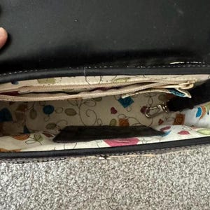 May include: Open black leather handbag with a patterned interior. The interior fabric features colorful floral and geometric designs. The bag has multiple compartments and a silver-colored clasp.