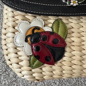May include: A woven straw handbag with a black leather top. The bag features a decorative ladybug and flower applique. The ladybug is red with black spots, and the flower is white with a yellow center. Green leaves accent the design.