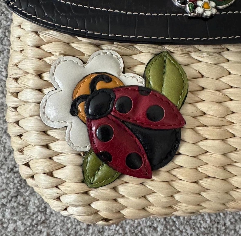 May include: A woven straw handbag with a black leather top. The bag features a decorative ladybug and flower applique. The ladybug is red with black spots, and the flower is white with a yellow center. Green leaves accent the design.