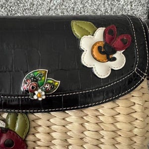 May include: Black crossbody bag with a woven straw base and a faux-leather flap. The flap features decorative ladybug and flower appliqu&eacute;s. The bag has a stitched border and a textured, crocodile-like pattern.