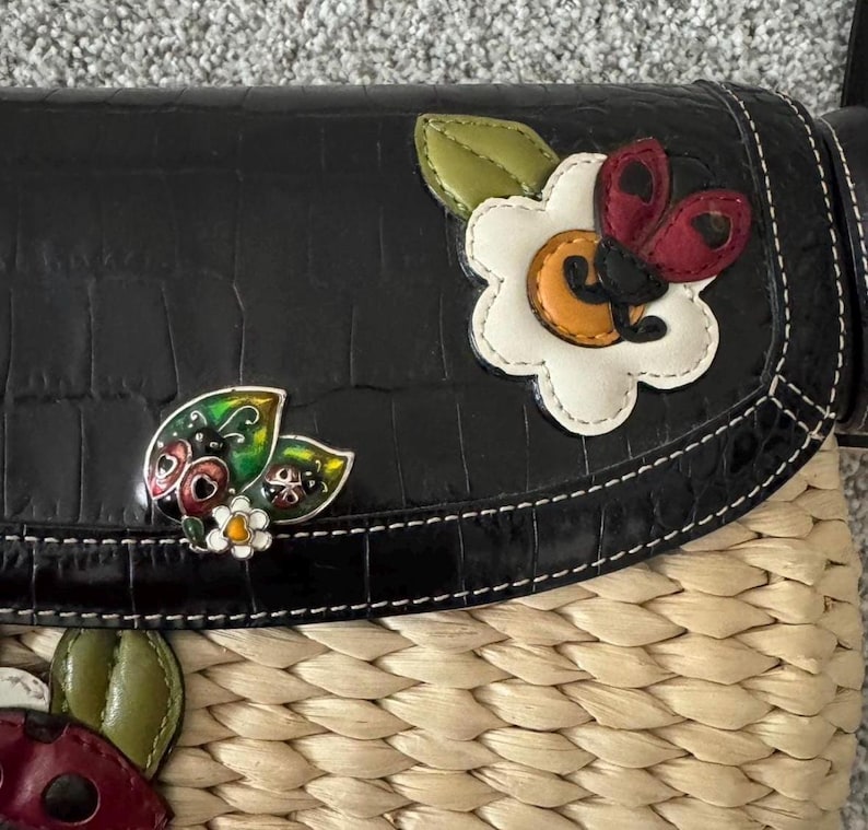 May include: Black crossbody bag with a woven straw base and a faux-leather flap. The flap features decorative ladybug and flower appliqu&eacute;s. The bag has a stitched border and a textured, crocodile-like pattern.