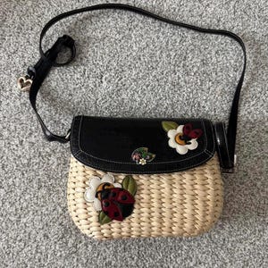 May include: A woven straw crossbody bag with a black flap and strap. The bag features decorative appliqu&eacute;s of a ladybug, flowers, and a butterfly. The strap has a heart-shaped charm.