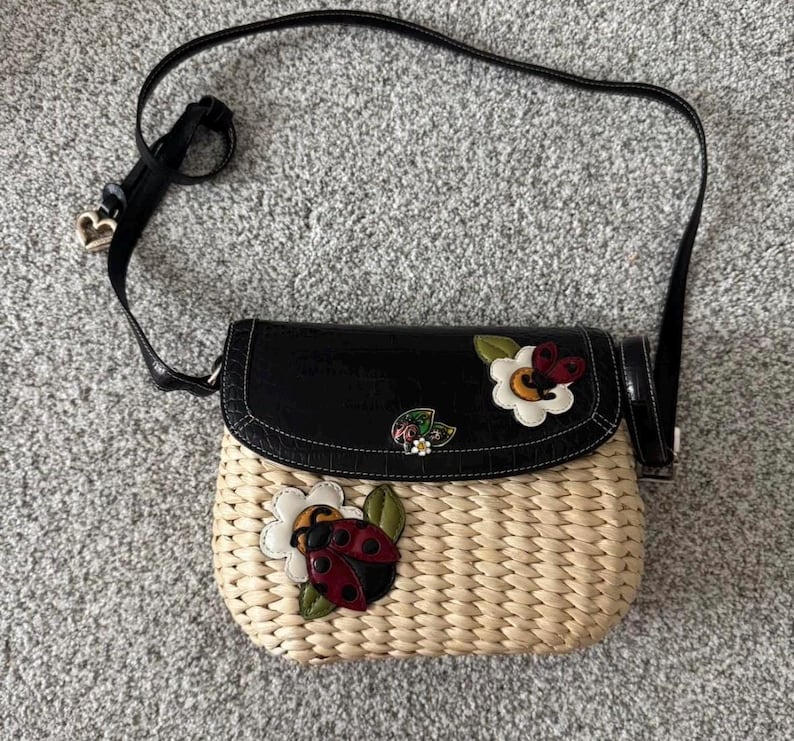 May include: A woven straw crossbody bag with a black flap and strap. The bag features decorative appliqu&eacute;s of a ladybug, flowers, and a butterfly. The strap has a heart-shaped charm.