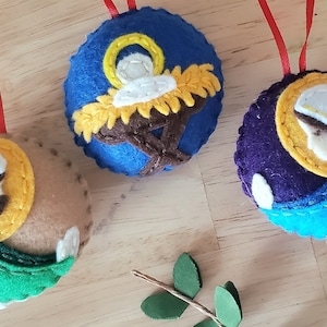 May include: Three felt Christmas decorations depicting scenes from the Nativity. The first decoration is blue with a yellow star and depicts the baby Jesus in a manger. The second decoration is brown with a yellow halo and depicts Mary holding the baby Jesus. The third decoration is purple with a blue and white halo and depicts Joseph.
