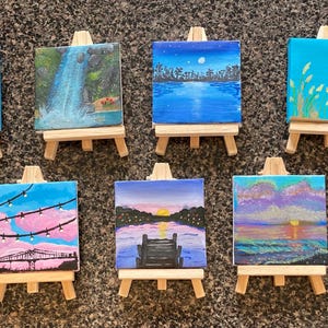 Könnte beinhalten: Zehn kleine Leinwände auf Staffeleien, jede mit einer anderen Szene bemalt. Die Szenen umfassen einen Wasserfall, einen Leuchtturm, eine Brücke, einen Sonnenuntergang und eine Stadtlandschaft. Die Leinwände sind alle in leuchtenden, lebendigen Farben bemalt.