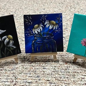 Könnte beinhalten: Drei kleine quadratische Gemälde auf Holzstaffeleien. Eines zeigt weiße Blumen auf schwarzem Hintergrund, ein anderes Gänseblümchen in einem blauen Glas und das dritte eine rosa Blume auf türkisfarbenem Hintergrund. Jedes Gemälde ist etwa 10 cm groß.