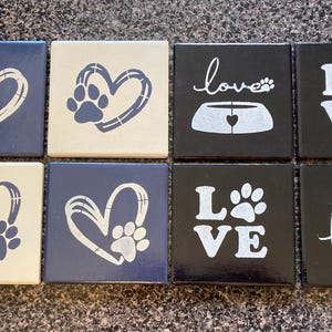 Könnte beinhalten: Acht quadratische Untersetzer aus Keramik mit verschiedenen Designs. Die Designs beinhalten Herzen, Pfotenabdrücke, das Wort "love" und eine Hunde-Schüssel. Die Untersetzer sind in Schwarz, Weiß und Blau bemalt.