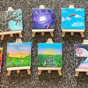 Könnte beinhalten: Zehn kleine Leinwandbilder auf Staffeleien, die jeweils eine andere Szene darstellen. Die Bilder zeigen einen Regenbogen, einen Sonnenuntergang, einen Mond, einen Baum und ein Herz. Die Bilder sind alle in leuchtenden Farben gehalten und sehr detailliert.