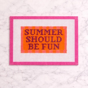 May include: A rectangular needlepoint canvas with a bright pink border and a white center. The center features the text "SUMMER SHOULD BE FUN" in purple letters on an orange background. The canvas is on a marble surface.