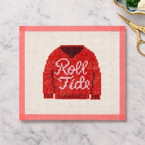 May include: A needlepoint design featuring a red sweater with the words "Roll Tide" in white script. The design is on a white canvas with a pink border. The needlepoint is likely for a decorative item or a craft project.